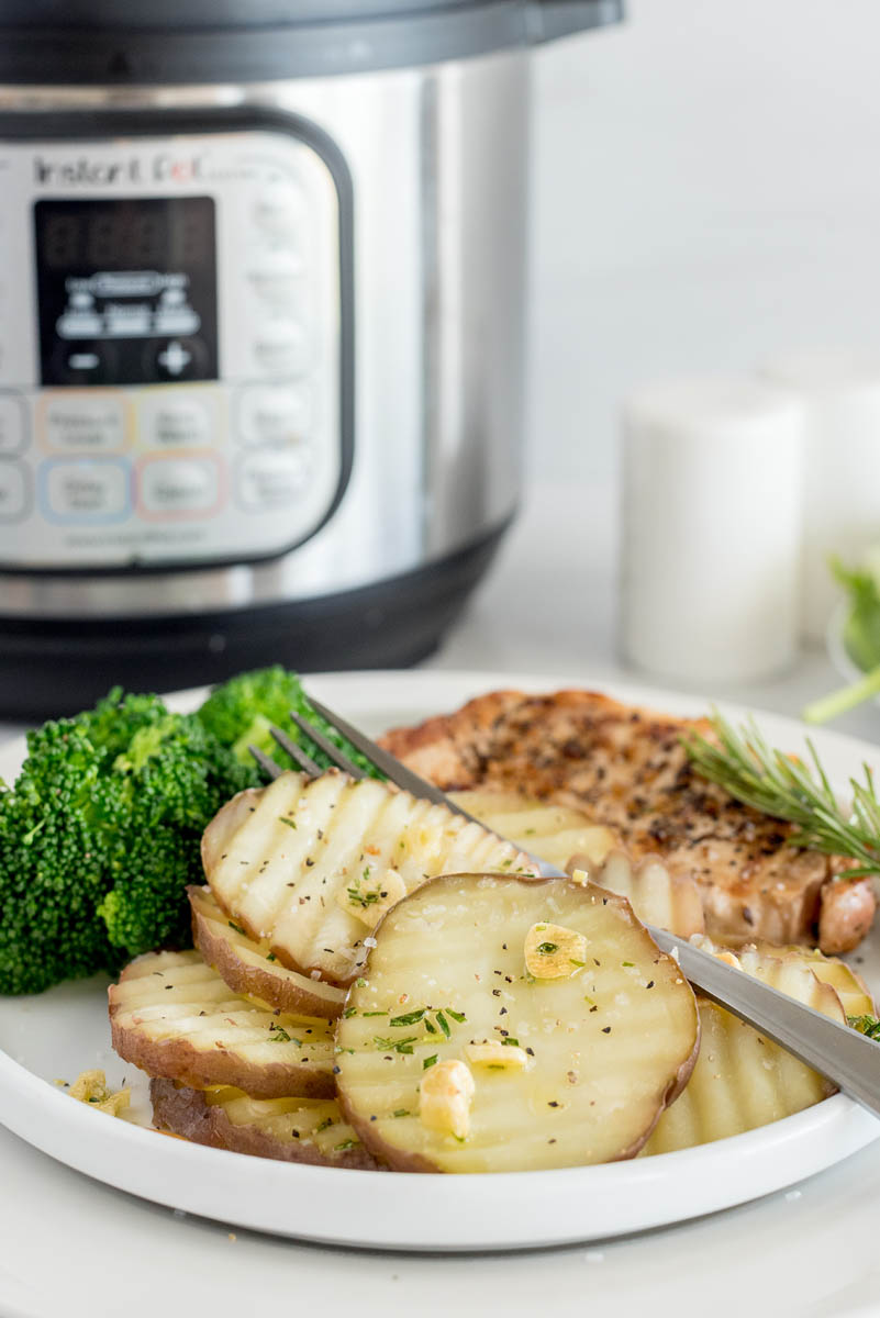 A 45-degreee shot of a white plate of rosemary new potatoes with a silver fork resting on top, with an out of focus Instant Pot in the background