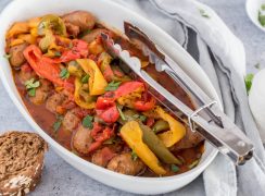 A white serving dish filled with Instant Pot sausage and peppers with tongs for serving, placed next to a gray napkin.