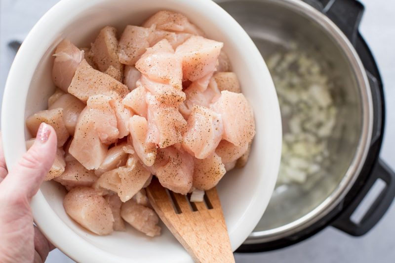 Overhead of raw diced chicken thighs seasoned with freshly ground black pepper being put into an Instant Pot with diced onions being sautéed for Pressure Cooker Honey Sesame Chicken.