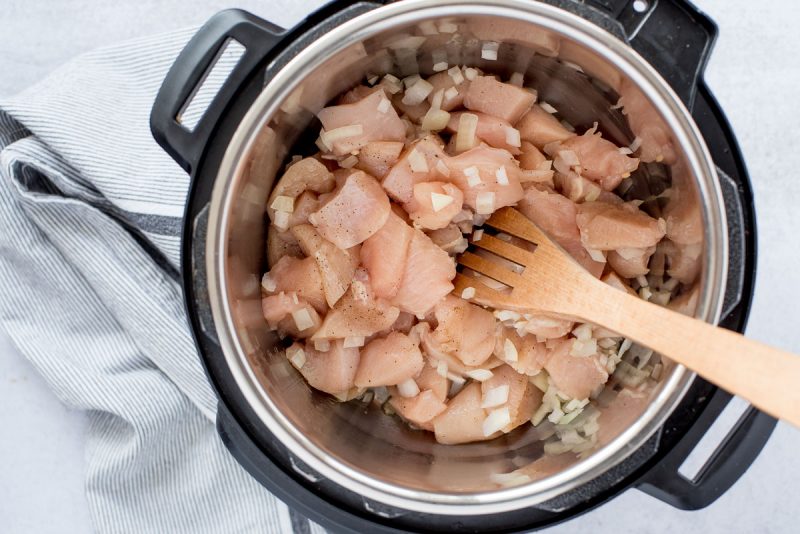 Overhead of an Instant Pot filled with diced chicken thighs seasoned with black pepper being sautéed with diced onion, garlic, and olive oil and stirred with a wooden spoon to make Pressure Cooker Honey Sesame Chicken.