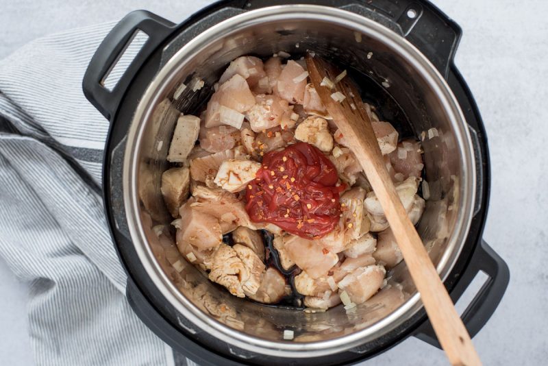 Overhead of pressure-cooked diced chicken thighs with ketchup, and red pepper flakes being stirred with a wooden spoon to make Instant Pot Honey Sesame Chicken.