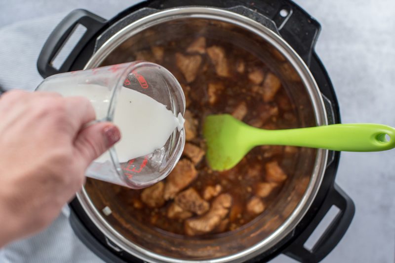 Overhead of a person using a liquid measuring cup to pour a cornstarch slurry into Instant Pot Honey Sesame Chicken in an electric pressure cooking being stirred with a green rubber spatula.