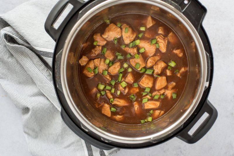 Overhead of an Instant Pot filled with cooked Honey Sesame Chicken garnished with fresh green scallions and made with diced chicken thighs and sauce flavored with soy sauce, honey, ketchup and thickened with a cornstarch slurry.
