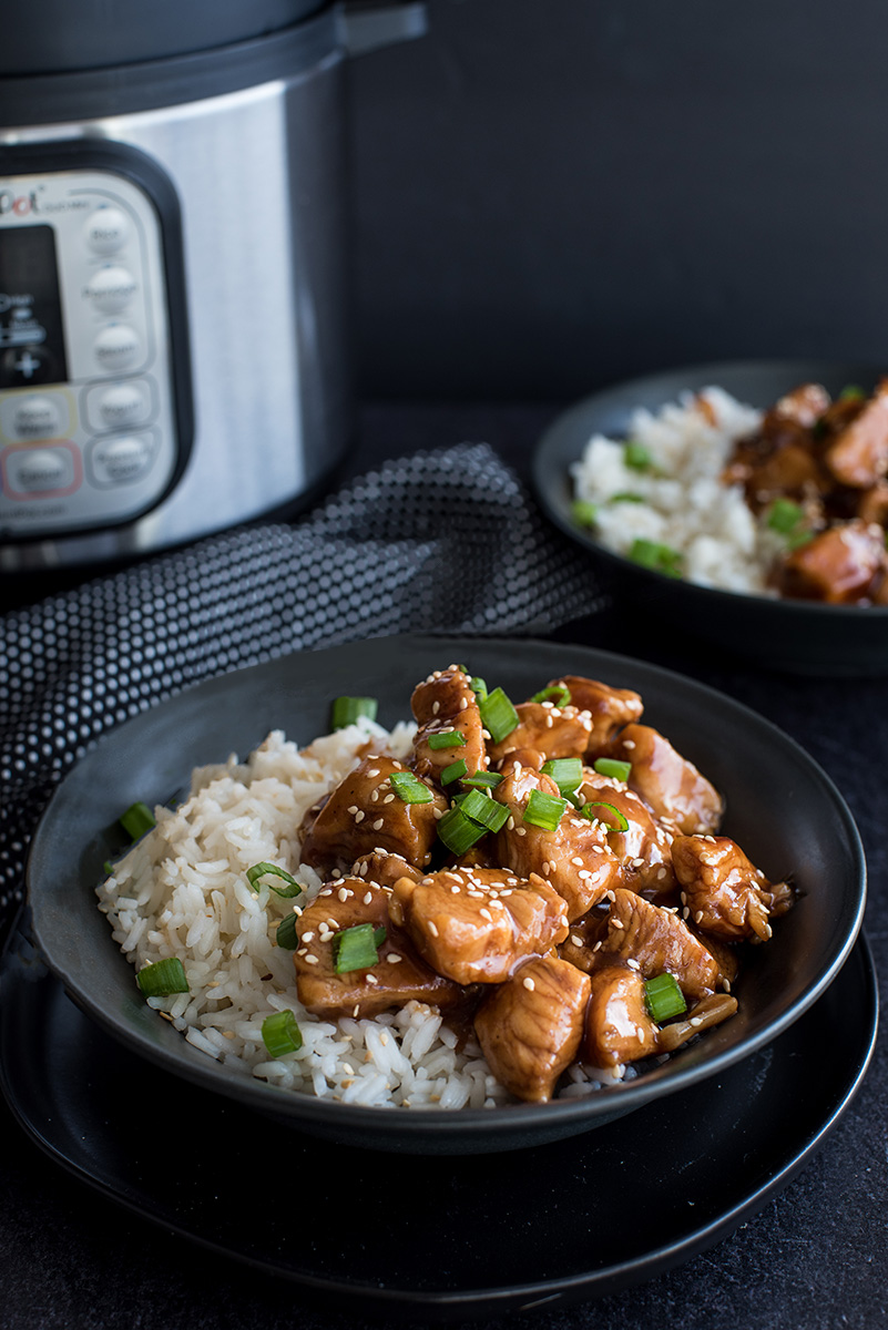 Close up on a black bowl filled with Instant Pot fluffy white rice and pressure cooker Honey Sesame Chicken garnished with sliced fresh scallions, white sesame seeds and sticky soy honey sauce with an Instant Pot in the background.