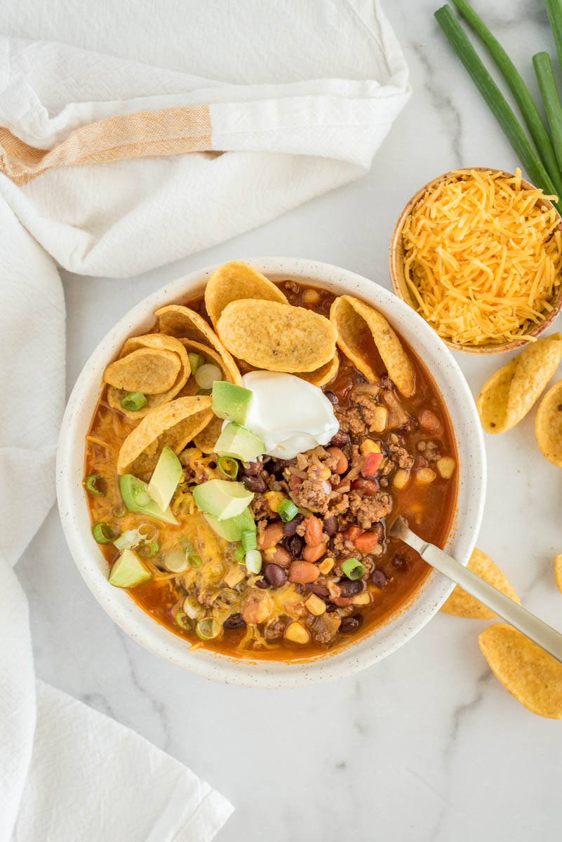 Overhead picture of Instant Pot taco soup topped with sour cream, cheese, avocado, and chips with shredded cheese and green onion in the background.