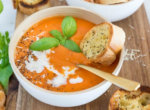 a 45 degree shot of a white bowl against a wooden background. The bowl is filled with orange tomato soup, garnished with basil, salt and pepper, and heavy cream, with a silver spoon and a baguette on the side.