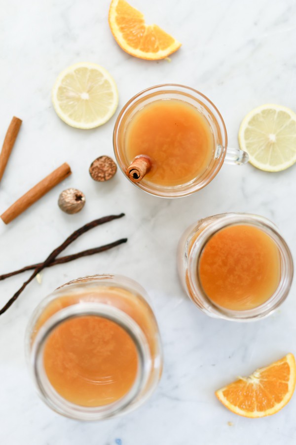 overhead photo of 2 mugs and a pitcher of wassail, surrounded by fresh orange and lemon slices, cinnamon sticks, and vanilla beans