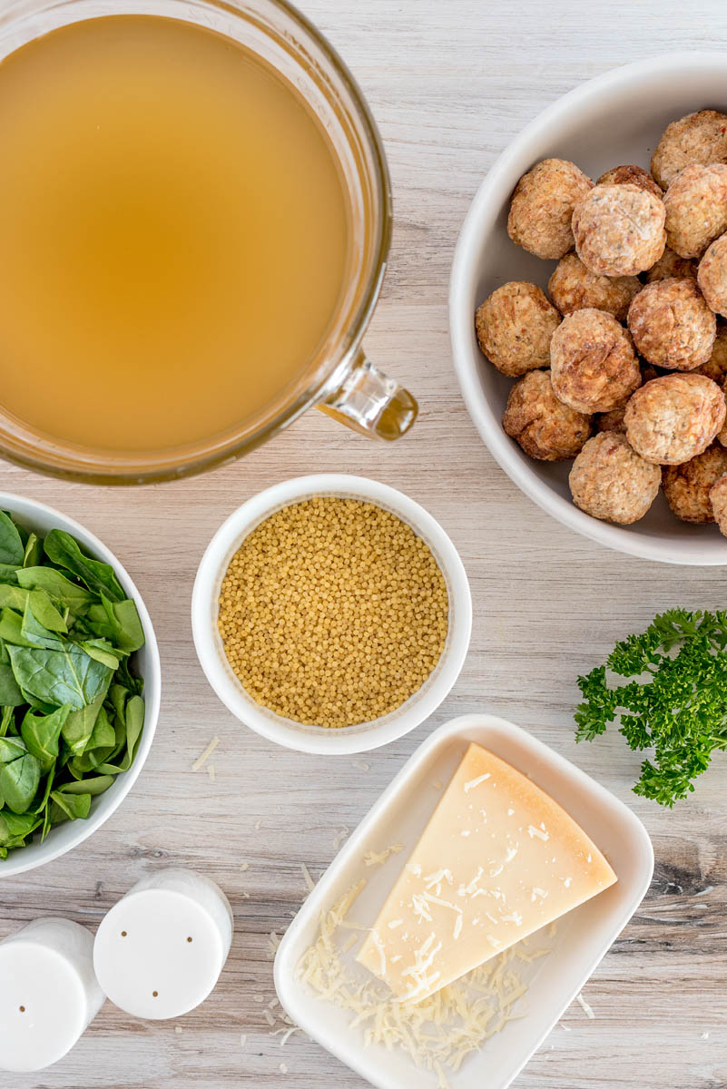 a vertical overhead shot of all the ingredients needed to make the Italian Wedding Soup recipe., including broth, spinach, ancini di pepe pasta, frozen meatballs, and parmesan cheese