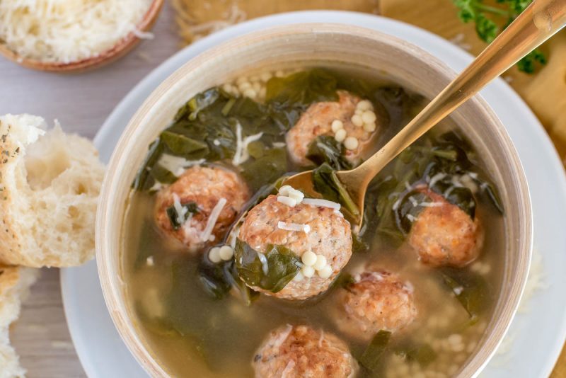 Overhead picture of a bowl of Instant Pot Italian wedding soup in a white bowl, with a spoon in the bowl, and bread and parmesan cheese in the background.